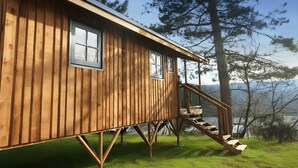 Una cabaña de madera con una escalera que lleva a una terraza, rodeada de árboles y vista al lago.