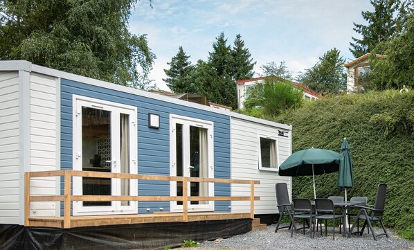 A mobile home with a deck, outdoor seating, and a covered patio area. - The Grande Large mobile home is a fully equipped, comfortable accommodation for 6 people, ideal in any season thanks to its three bedrooms, terrace, air conditioning, and central heating. (Dochamps)