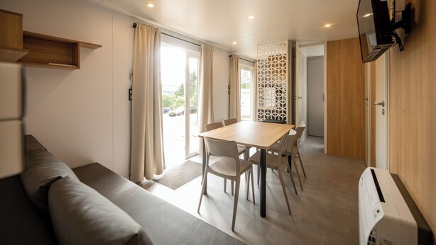 A modern room with a wooden dining table, a sofa, and a TV mounted on the wall. - The Grande Large mobile home is a fully equipped, comfortable accommodation for 6 people, ideal in any season thanks to its three bedrooms, terrace, air conditioning, and central heating. (Dochamps)