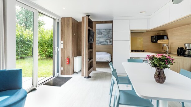 Quarto de hotel moderno com uma grande porta de vidro deslizante para um jardim, área de jantar com mesa branca e cadeiras azuis, cozinha compacta e cama com cabeceira branca.