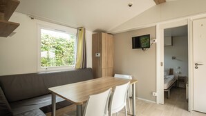 Quarto com mesa de madeira, cadeiras brancas, sofá cinza, televisão fixada na parede e porta que dá para outro cômodo.