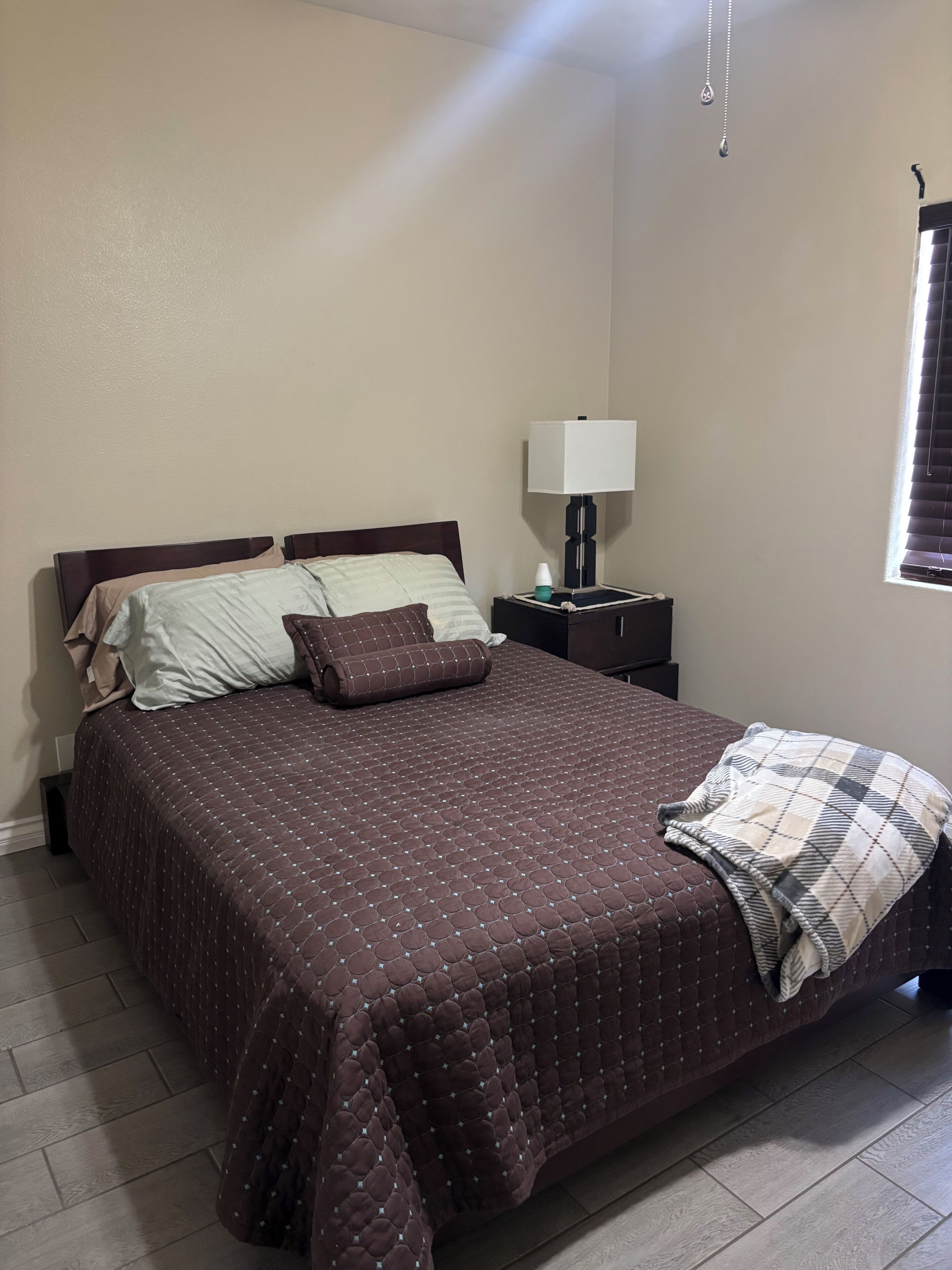 A neatly made bed with a brown quilt, a bedside lamp, and a window with blinds.