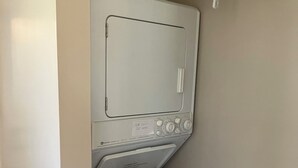 A compact, white, front-loading washer and dryer unit with a control panel, a caution label, and a water inlet hose. - Comfortable 1-bedroom apartment close to Beach with AC- Vero Beach (Vero Beach)