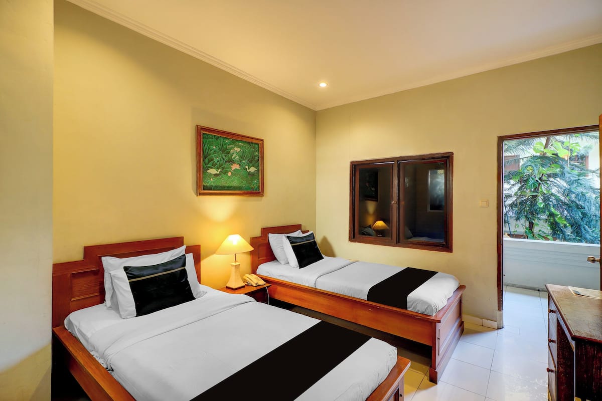 A hotel room with two beds, a nightstand with a lamp, a wooden headboard, a framed picture on the wall, and a window with a view of greenery.