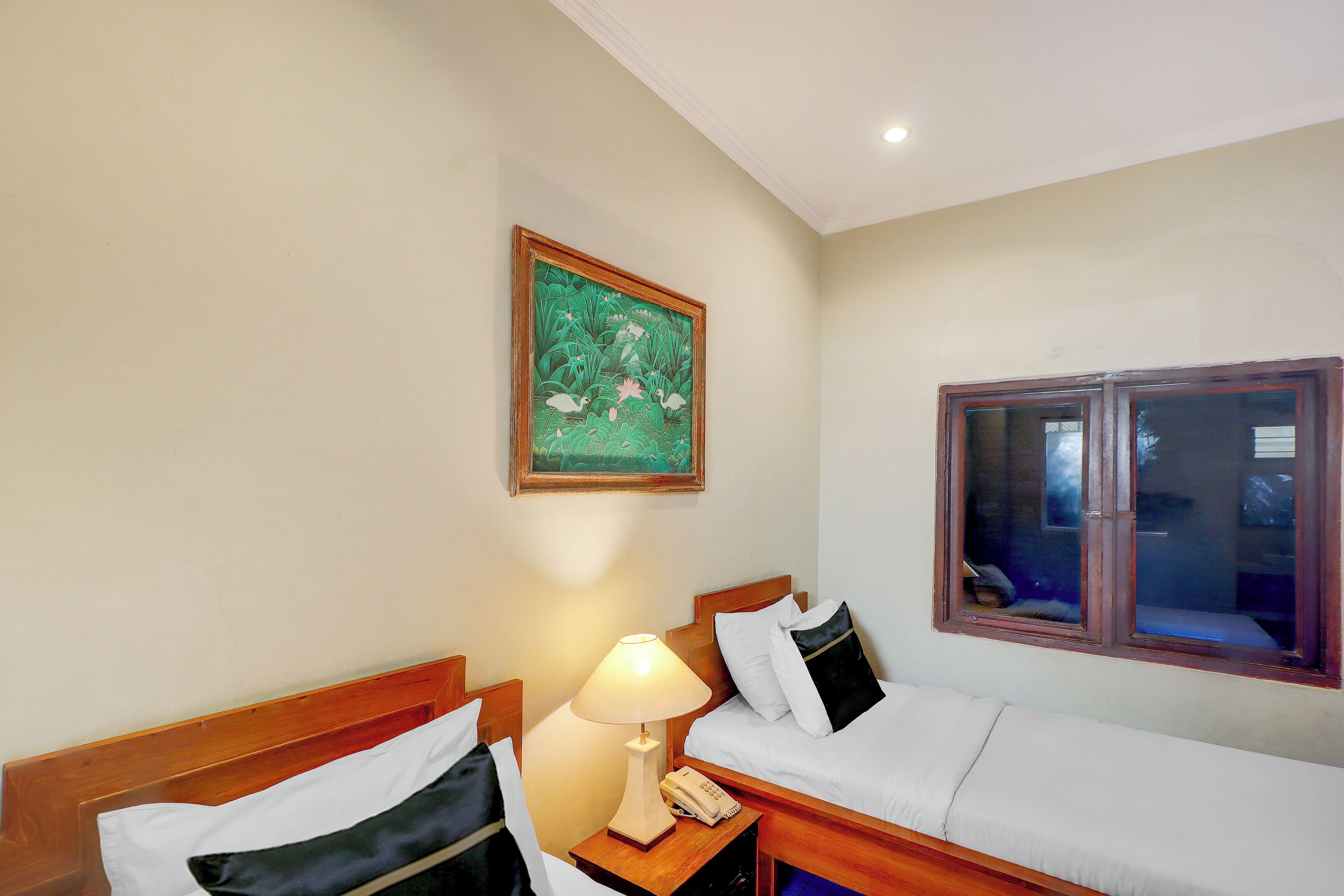 A hotel room with two beds, a nightstand with a lamp, a telephone, a framed painting on the wall, and a window with a view.