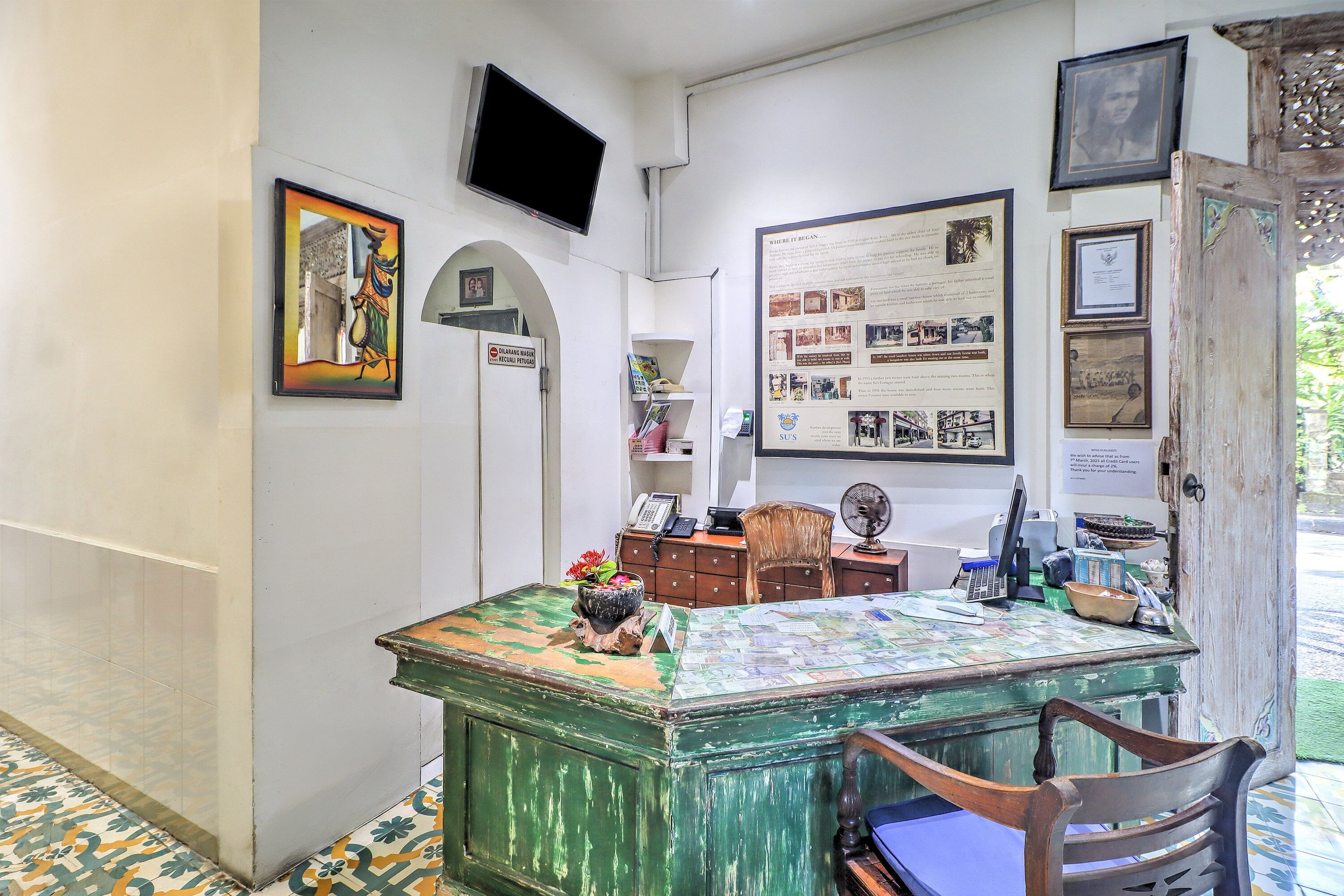 Una habitación con un escritorio verde, una silla, un televisor montado en la pared y un tablón de anuncios con diversas imágenes y texto.