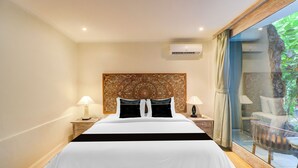A hotel room with a large bed, two bedside tables with lamps, an air conditioning unit, and a view of greenery through a glass door. - Townhouse Oak Su's Cottage Near Legian Beach (Kuta)