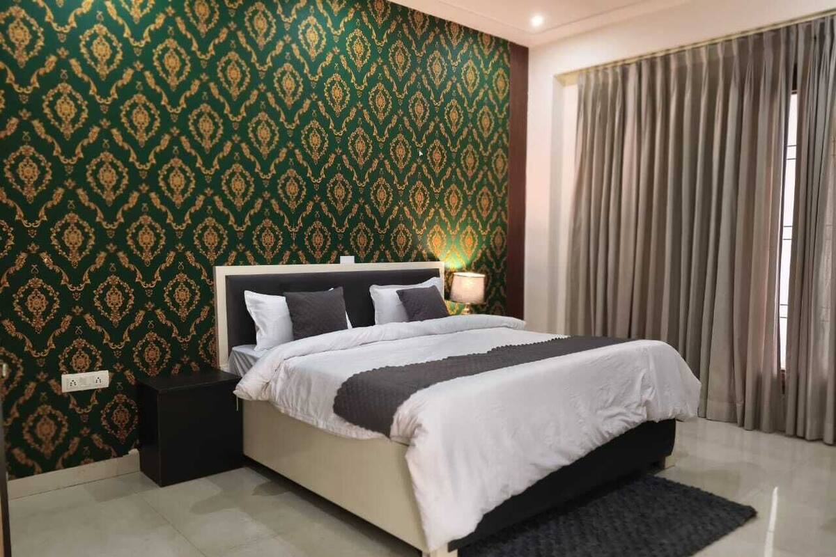 Un dormitorio con cama, mesita de noche, lámpara, alfombra oscura y pared con estampado.