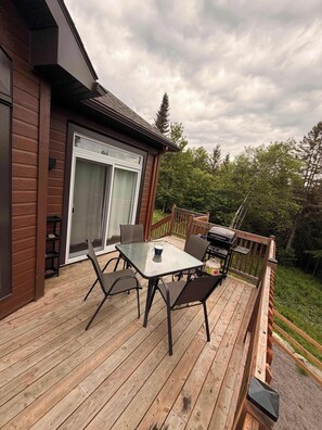 Una terraza de madera con mesa y sillas, una puerta corrediza de vidrio y una parrilla.