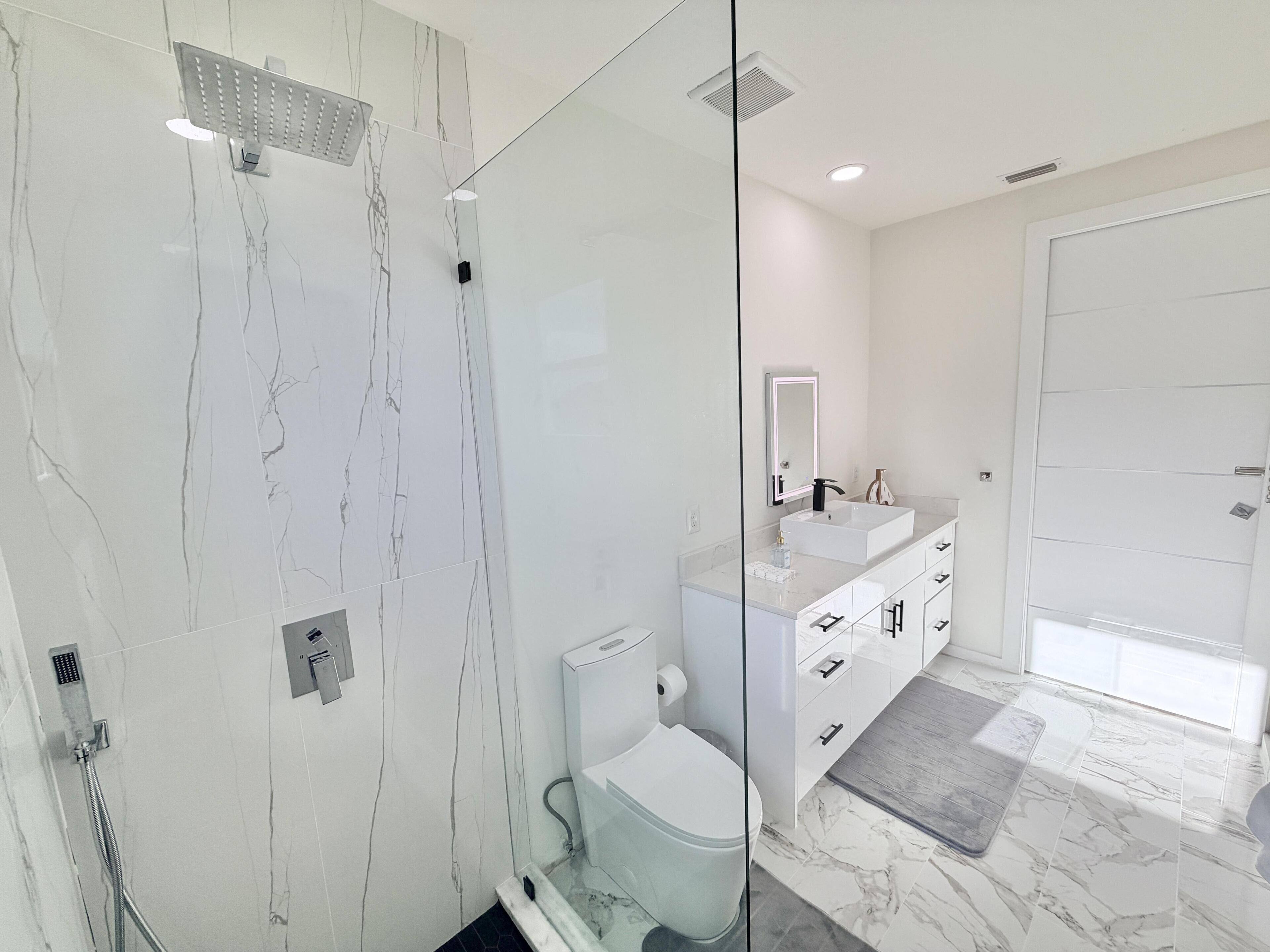 Un baño moderno con ducha envuelta en vidrio, inodoro blanco, tocador con espejo y un espejo grande sobre el lavamanos.