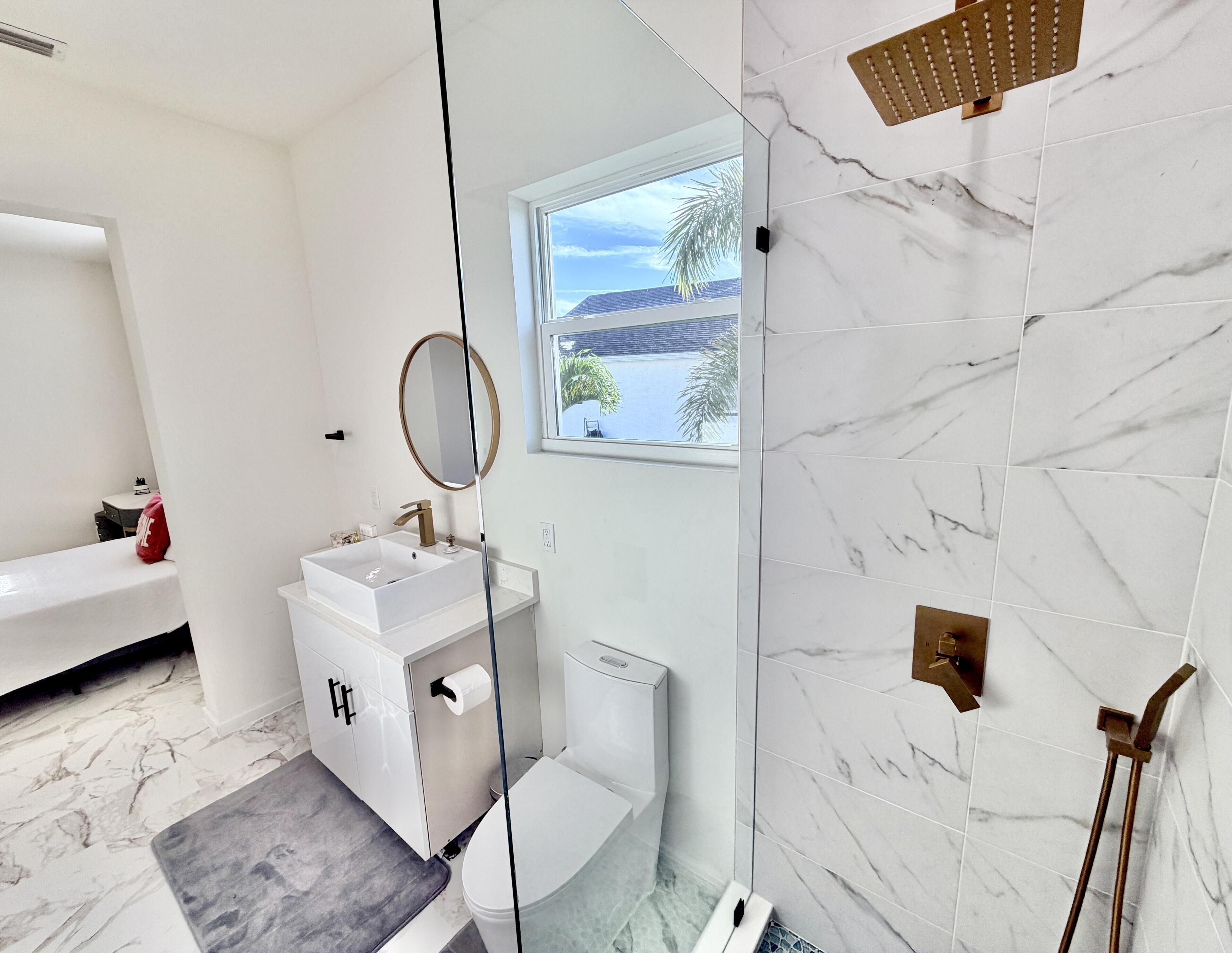 A modern bathroom with a white sink, a toilet, a glass shower enclosure, and a window with a view of the outdoors.