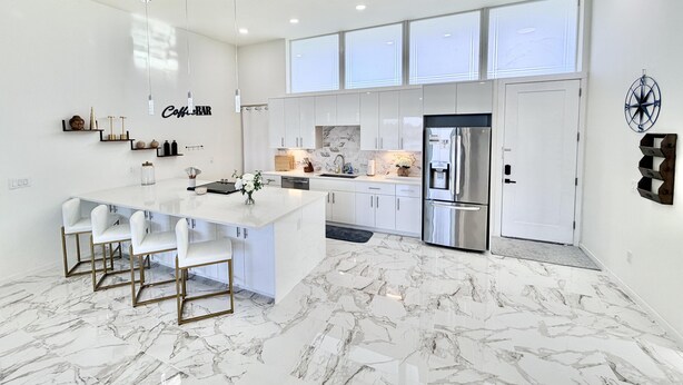 Una cocina moderna con una isla central, muebles blancos y un refrigerador de acero inoxidable.