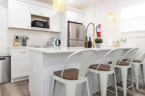 A modern kitchen with white cabinetry, a stainless steel refrigerator, a coffee maker, and a sink with a hose. There are three white chairs with wooden seats around the island. - Coastal Charm, 1st floor, AC, KingB, Free parking (Wildwood Crest)