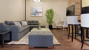 Uma sala moderna com sofá seccional, mesa de centro, área de jantar com cadeiras, planta em vaso e um quadro na parede.