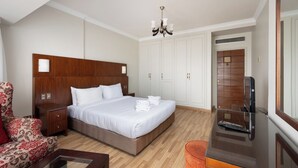A hotel room with a wooden headboard bed, white linens, a red patterned armchair, a small table, a television, and a wardrobe. - VESTA  - Luxury APT - 4BR - Agouza (I) (Giza)