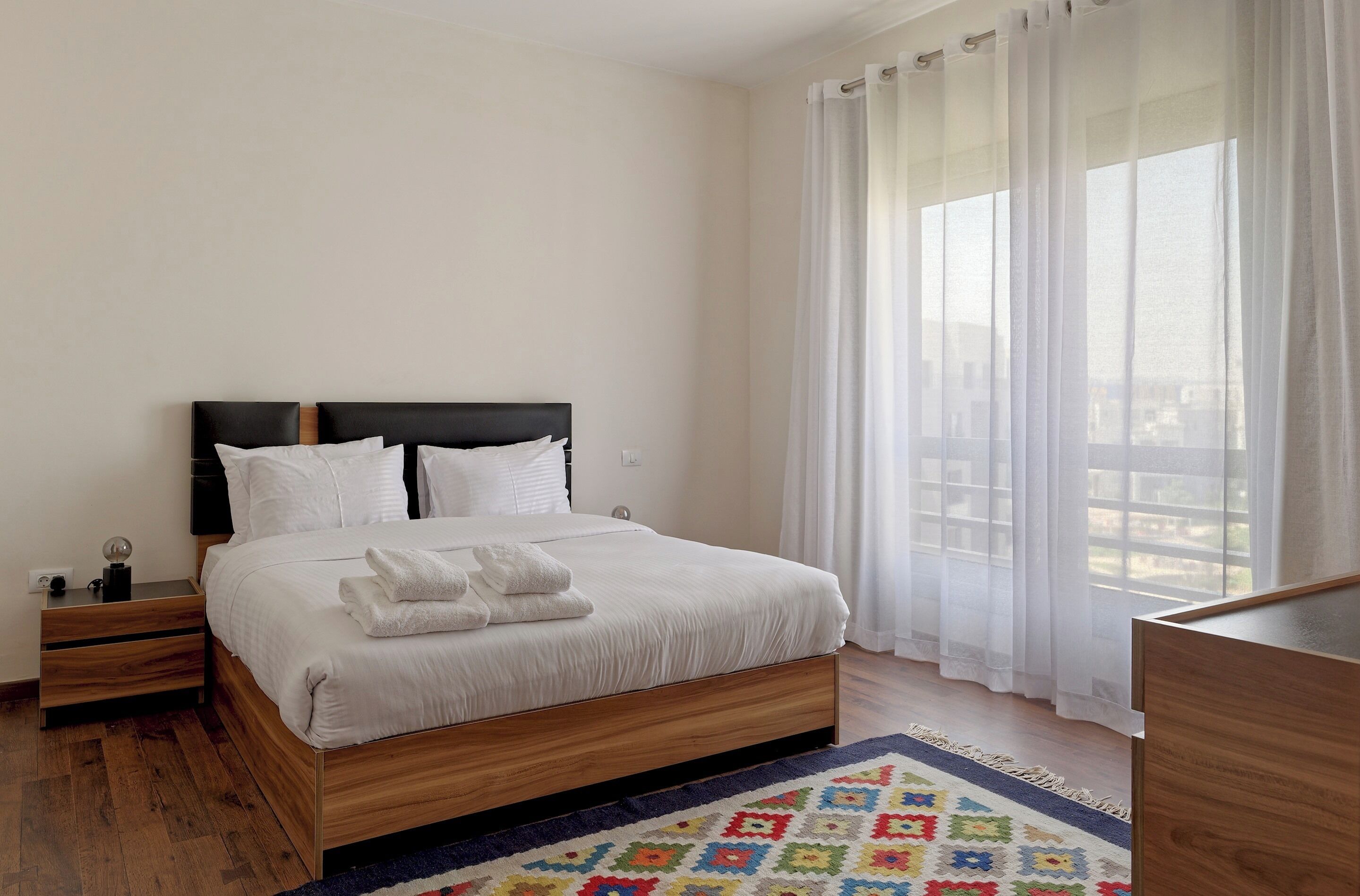 A neatly arranged hotel room with a bed, a nightstand, a desk, and a view through sheer curtains.