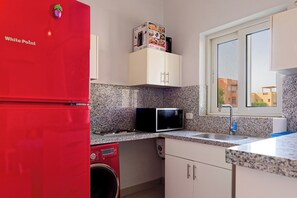 A compact kitchen with a red refrigerator, a microwave, a washing machine, and a window with a view of buildings.