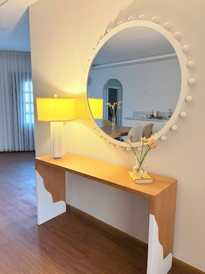 Een kamer met een houten console tafel, een ronde spiegel met verlichting, een lamp en een vaas met bloemen.