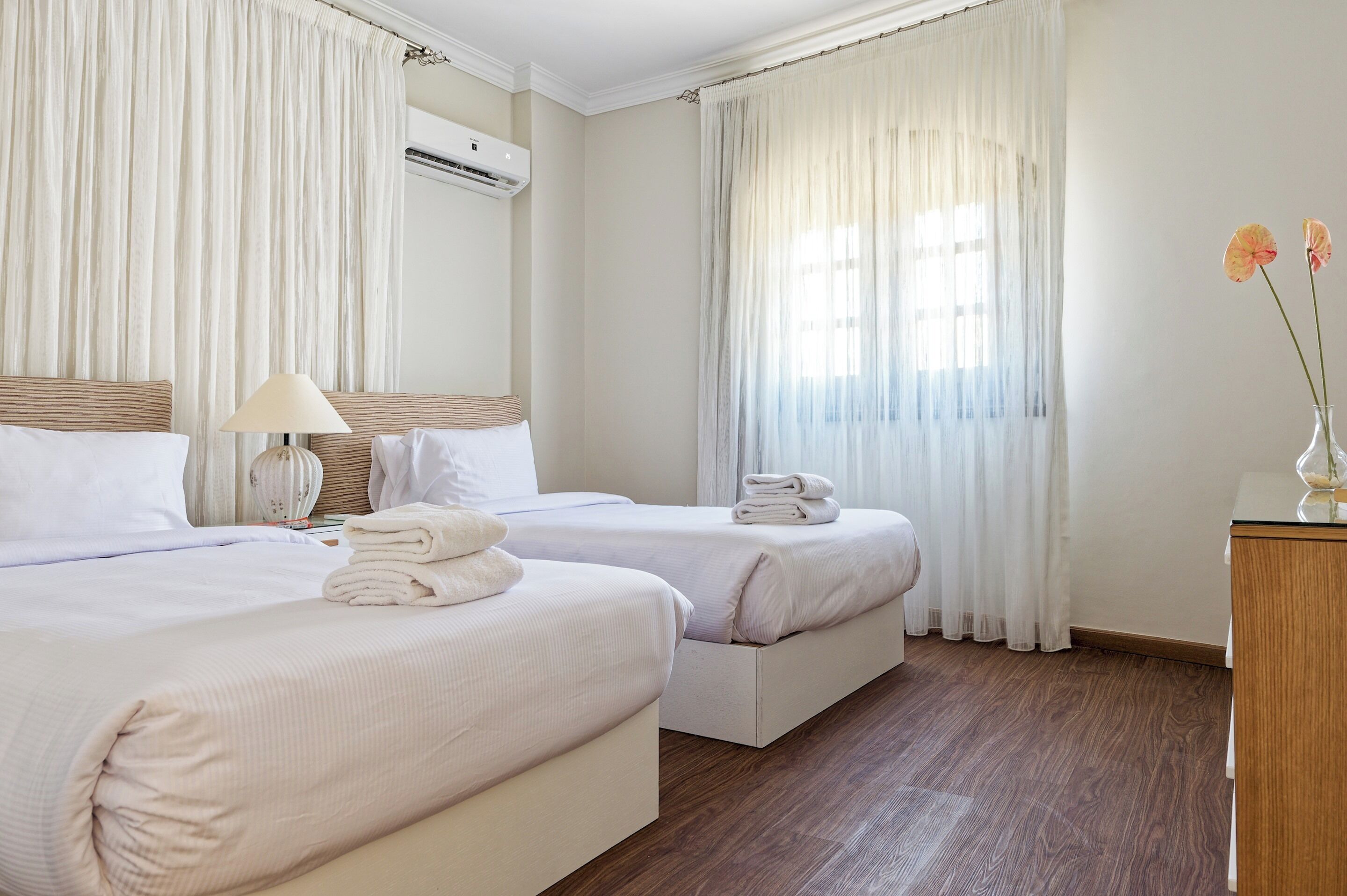 Une chambre d’hôtel avec deux lits, une table de chevet avec une lampe, un petit vase avec des fleurs et une commode en bois.