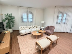Een woonkamer met een bank, fauteuil, salontafel en een kleine tafel met een lamp. Er staat een potplant en er is een flatscreen televisie.