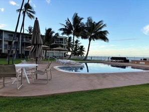 A pool area with lounge chairs, umbrellas, and palm trees. - Escape to Maili Cove in our fully furnished condo perfect for a family vacation. (Waianae)