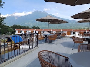 A terrace with tables and chairs, a view of a mountain, and a building with red-tiled roofs.