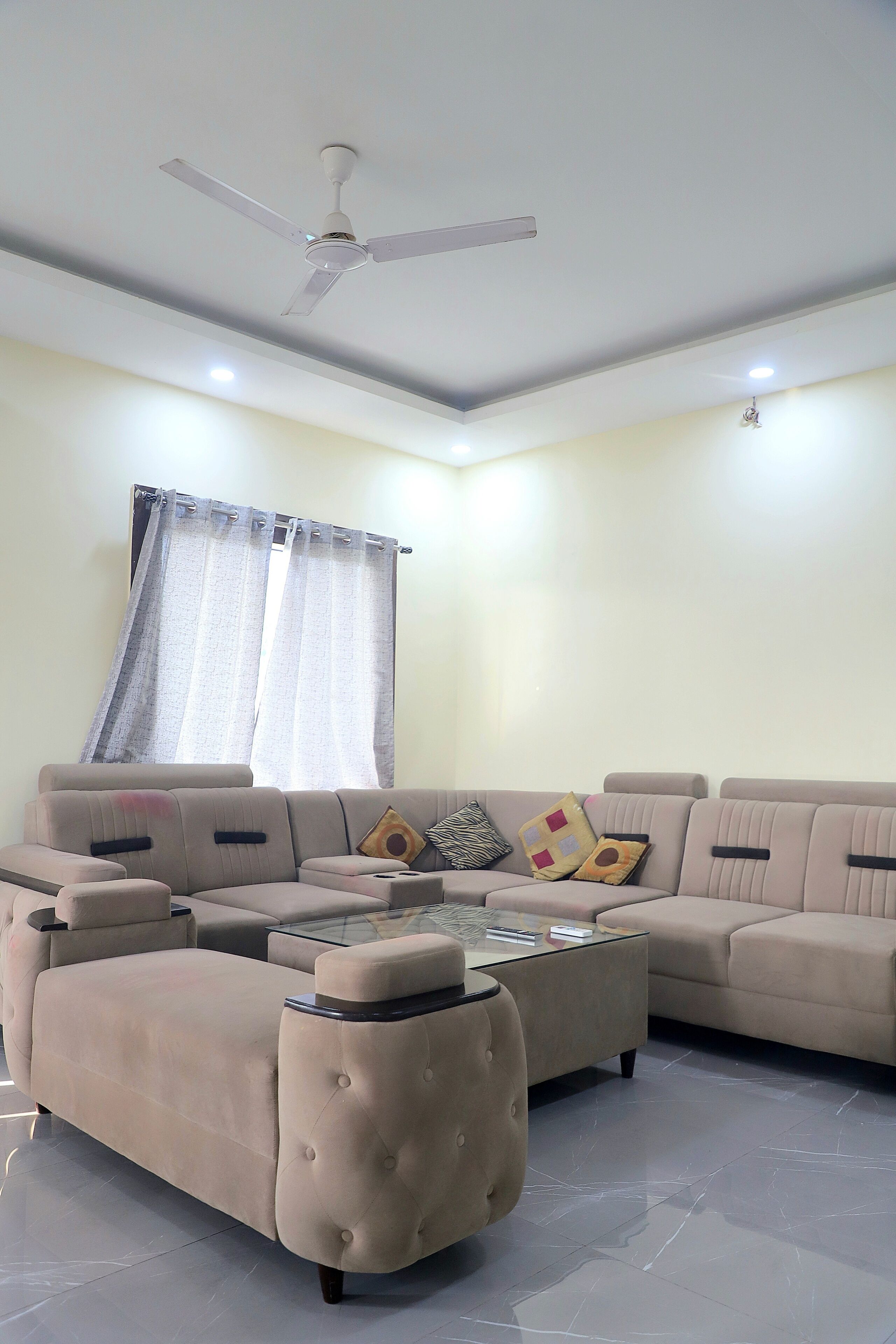Una sala de estar moderna con un sofá modular, una mesa de centro de vidrio, un ventilador de techo y una ventana con cortinas.