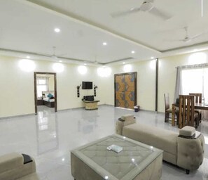 A spacious living area with a sofa, a coffee table, a TV, and a dining area with chairs and a table. - The Golden Farm (Noida)