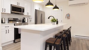 Una cocina moderna con armarios blancos, electrodomésticos de acero inoxidable, una isla central con taburetes y luces colgantes. Se observa una unidad de aire acondicionado.