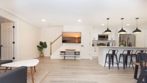 Una cocina moderna con una isla central, un área de comedor con taburetes de bar y una escalera que lleva a una sala de estar con un televisor grande.
