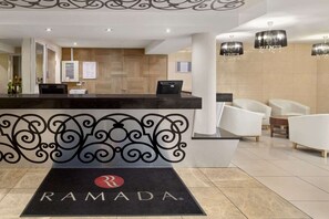 A reception area with a black reception desk, white chairs, and a black doormat with the logo 'RAMADA'. - Oldbury Birmingham (Oldbury)