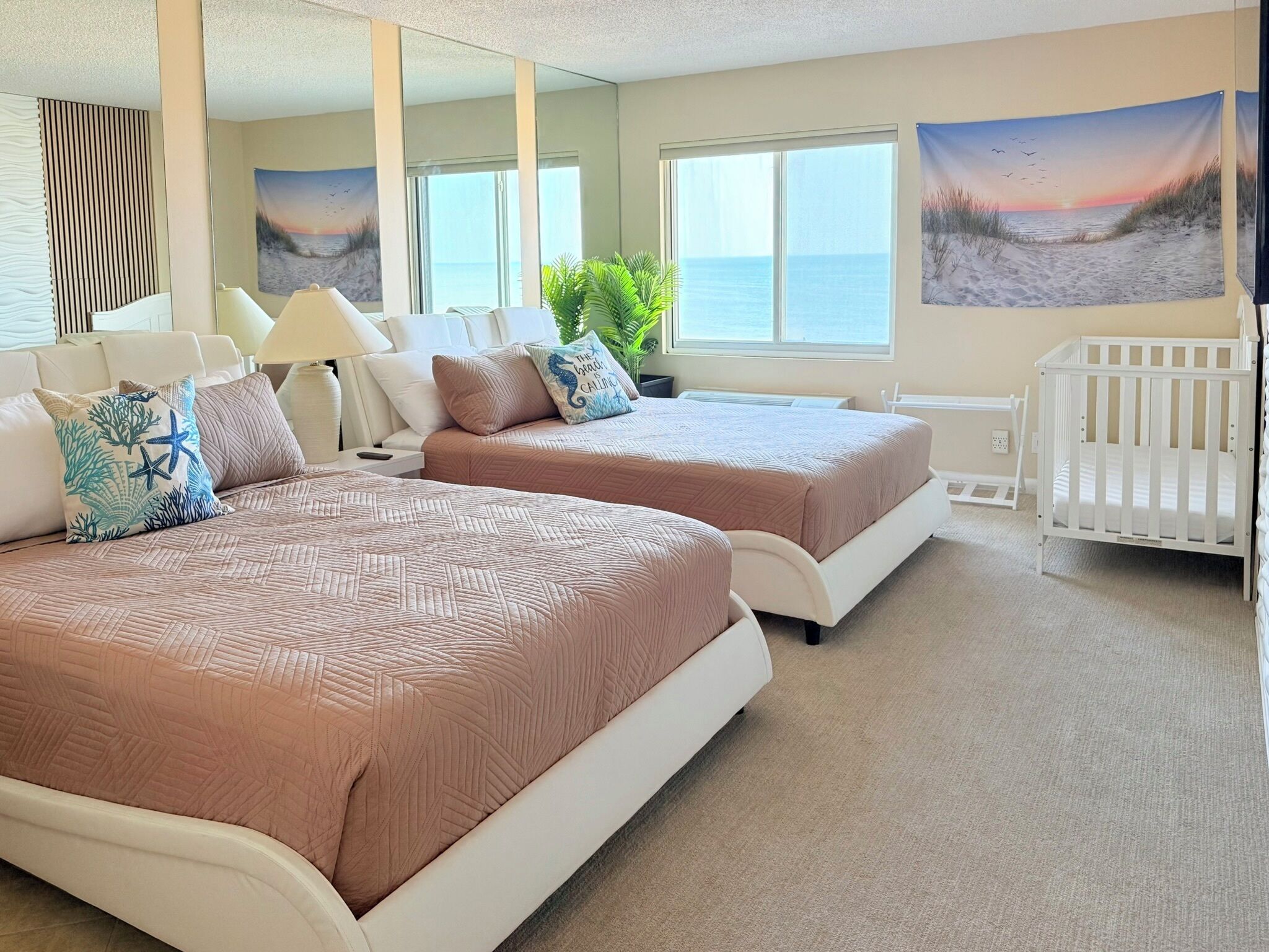 Een hotelkamer met twee bedden, een wiegje, grote ramen en strandafbeeldingen.