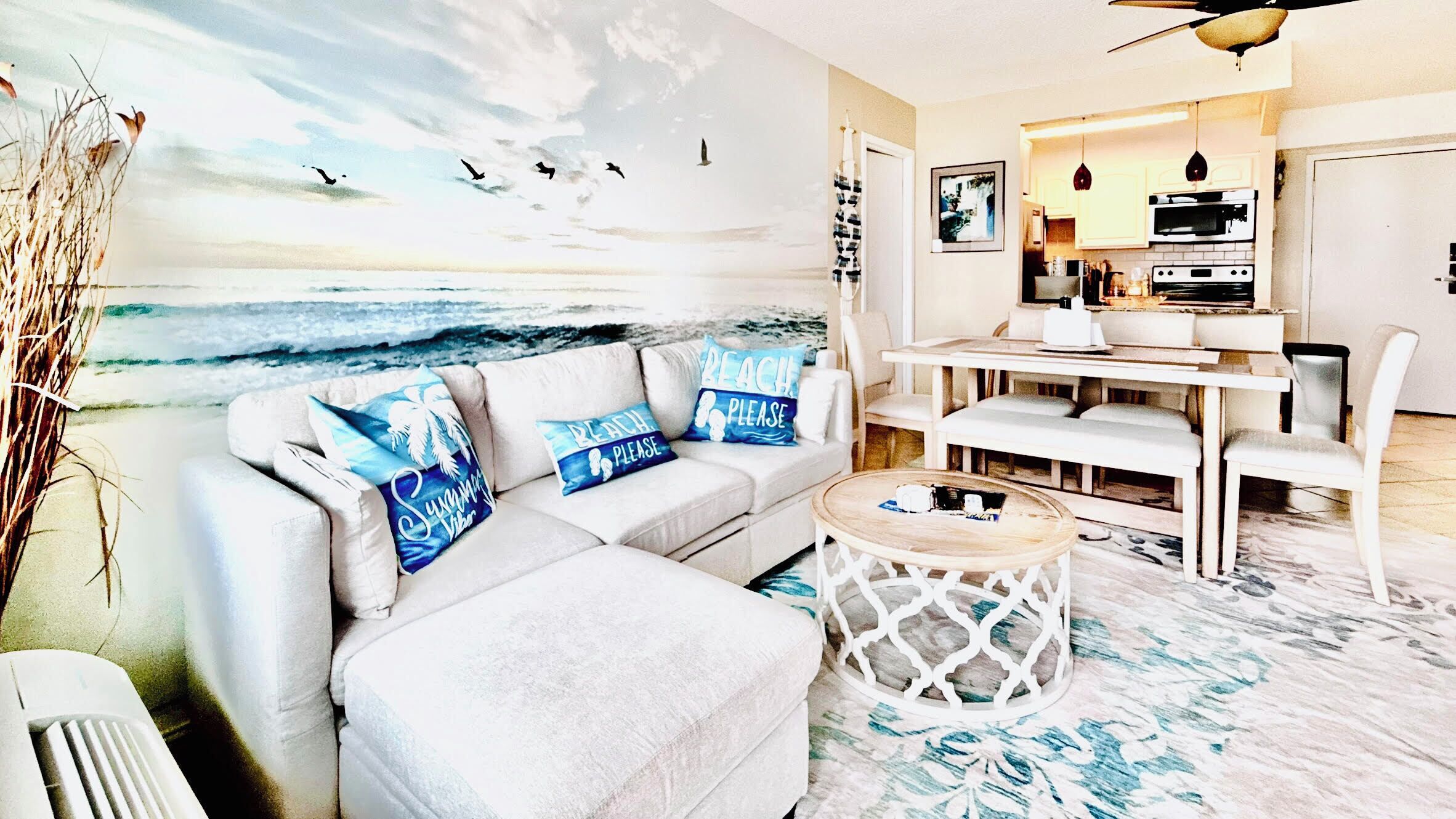 A living room with a beach mural, a sofa with beach-themed pillows, a dining area with a table and chairs, and a kitchen with appliances.