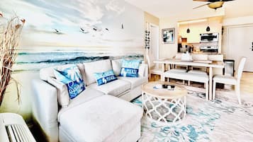 A living room with a beach mural, a sofa with beach-themed pillows, a dining area with a table and chairs, and a kitchen with appliances.