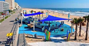 Un parque infantil en la playa con toldos azules, piscina y decoración con tablas de surf.
