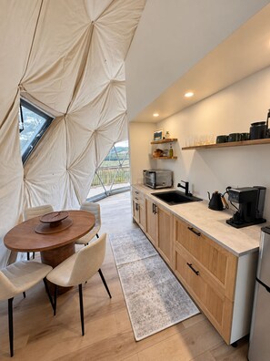 A modern kitchen with a dining area, featuring a round table, chairs, a coffee machine, and a view through a window. - The Flats - Premium Dome at Granda Rose Retreat (Branch)