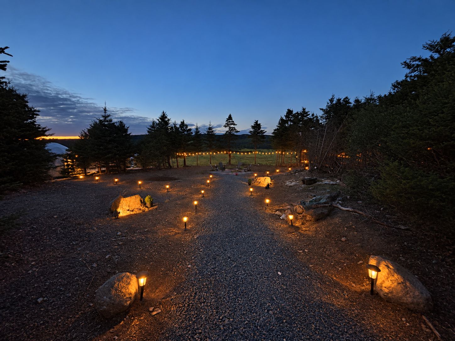 A gravel pathway with evenly spaced lanterns leading to a covered area with string lights.