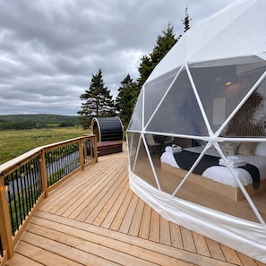 Estructura de domo geodésico con una cama en su interior, una terraza con barandilla y vistas a un paisaje natural.