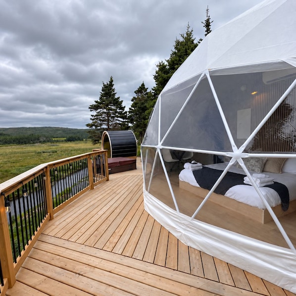 Eine geodätische Kuppel mit einem Bett im Inneren, einer Terrasse mit Geländer und Blick auf eine natürliche Landschaft.