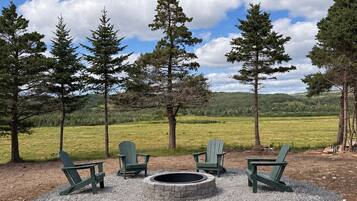 A gravel fire pit area with four Adirondack chairs, surrounded by trees and a grassy field under a partly cloudy sky.