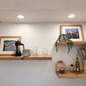 A kitchen with a coffee maker, glasses, a framed photo, a plant, and a bread loaf on a shelf.