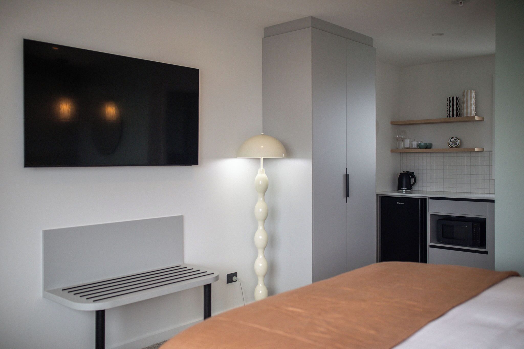 A modern hotel room with a bed, a bedside table, a lamp, a TV, a kitchenette, and a wardrobe.