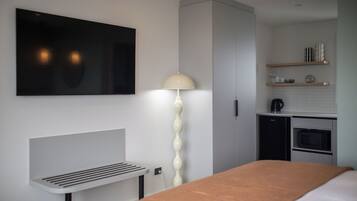 A modern hotel room with a bed, a bedside table, a lamp, a TV, a kitchenette, and a wardrobe.