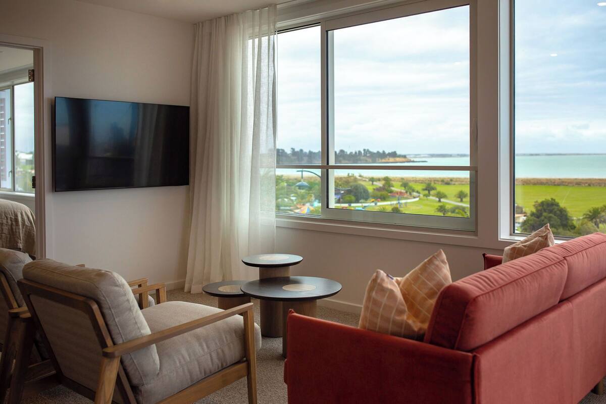 A living room with a TV, a sofa, a chair, and a view of a body of water through large windows.