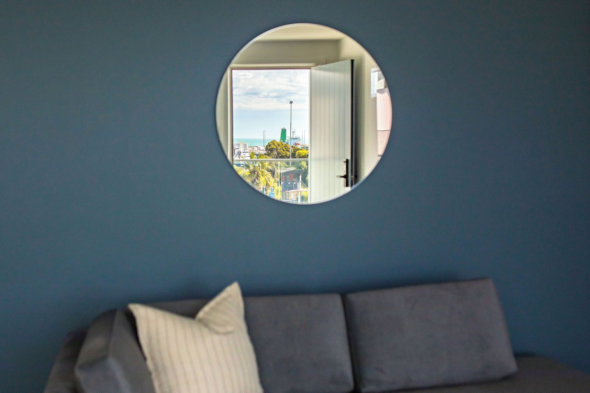 A circular mirror on a wall, a gray sofa with a pillow, and a view of a cityscape through the mirror.