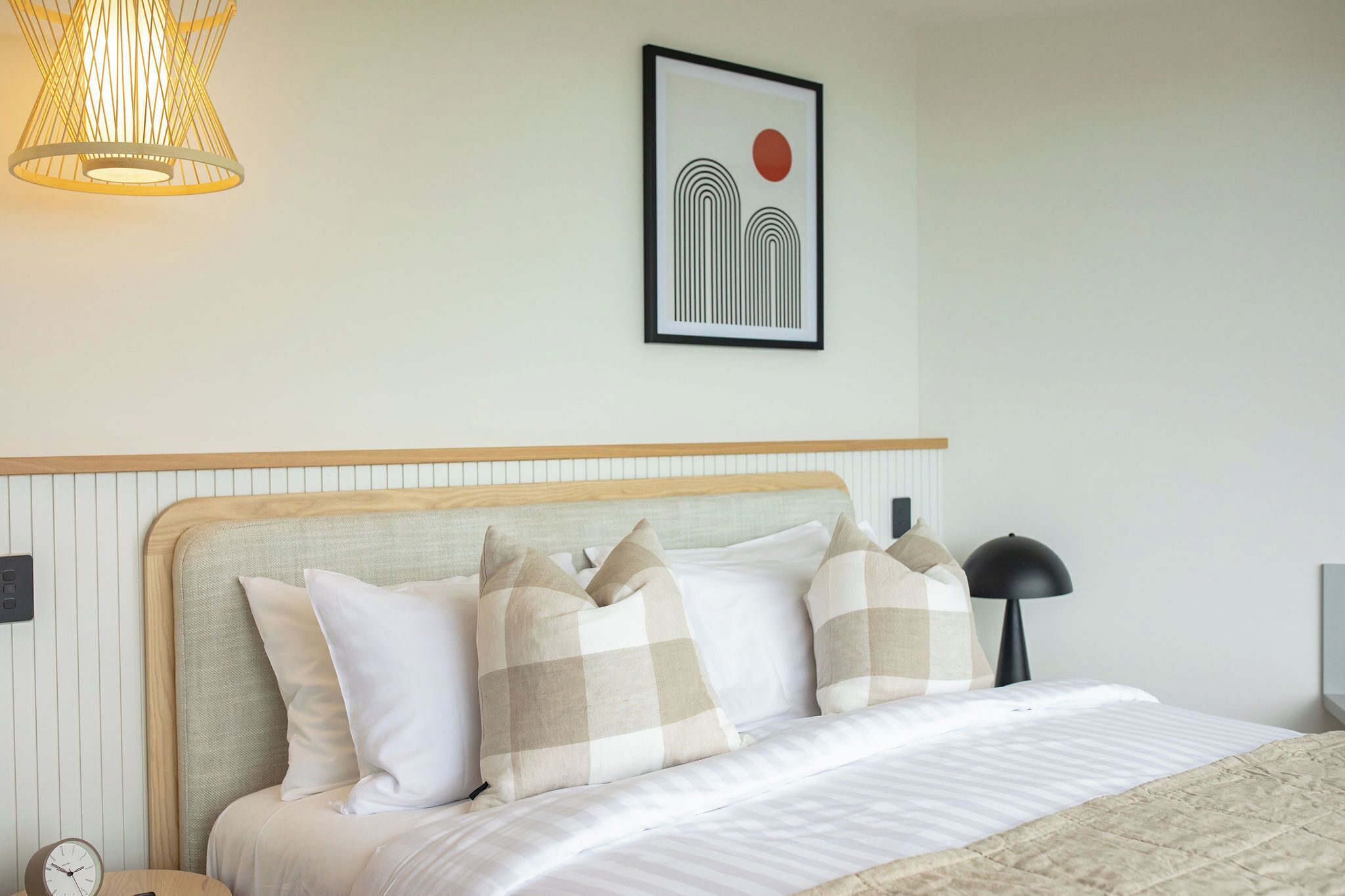 Una cama bien hecha con almohadones cuadriculados, una lámpara de noche, una obra de arte enmarcada en la pared y un colgador de luz.