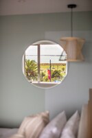 Una habitación con ventanal circular que da a una playa, una lámpara y una cama con almohadas.