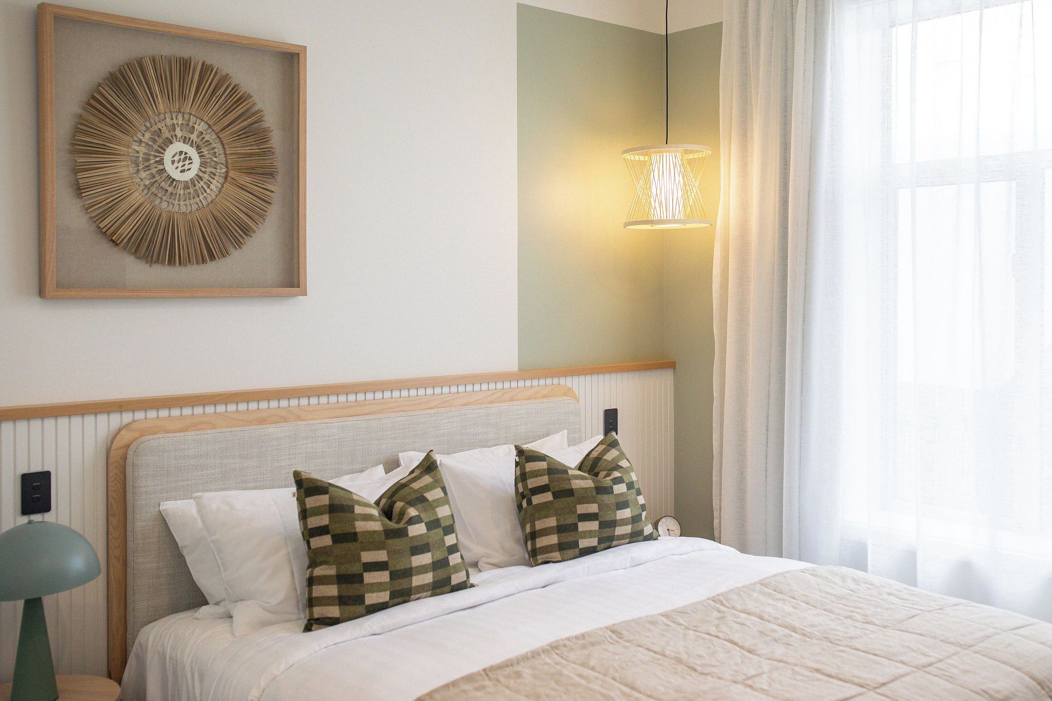 A neatly made bed with checkered pillows, a framed abstract artwork above it, and a bedside lamp.