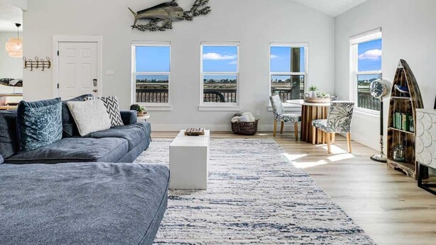 Ein geräumiges Wohnzimmer mit einem großen grauen Sofa, einem weißen Couchtisch und einem gemusterten Teppich. Drei Fenster bieten Ausblick ins Freie.