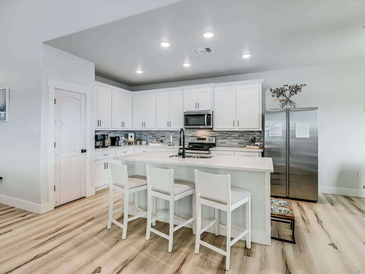 A modern kitchen with white cabinetry, a central island with bar stools, stainless steel appliances, and a wooden floor.